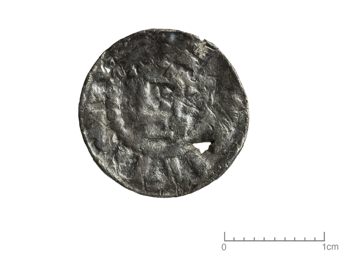 Mynt av sølv, Tyskland, av typen MgHP1 (Magdeburger Holzkirchenpfenninge). Adversen har deler av perlerad synlig mens tegn langs kanten og symbol i midten er nedslitt. Reversen har et kors i midten omgitt av en nedslitt perlerad samt tegn/bokstaver langs kanten. Mynten er slitt, bulket og det er et hull fra skade. Diameter er 1,7 cm og tykkelse 0,1 cm. 