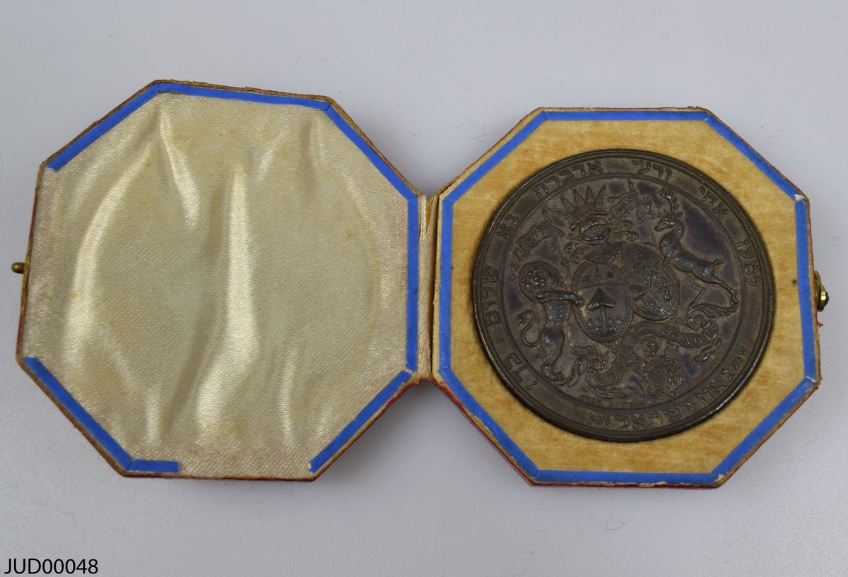 Medalj liggandes i pappask, som invändigt är klädd med siden och sammet. Medaljen är rikligt dekorerad med ett släktträd, vapensköld och hebreisk text på ena sidan samt flera namn på andra sidan.