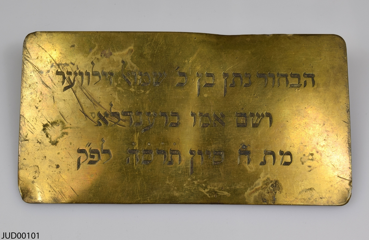 Två mässingsplattor liggandes i tygpåse med blomstermotiv. Plattorna är dekorerade med en ingraverad hebreisk text.