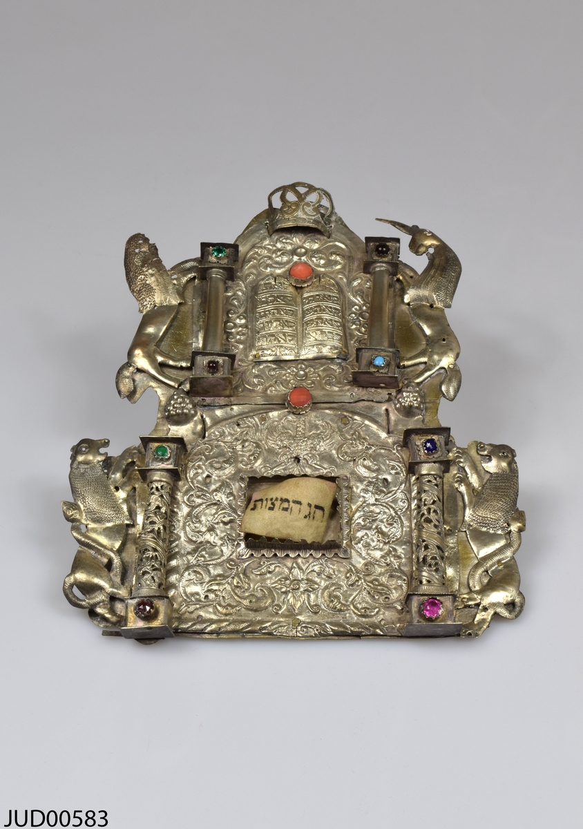 Torahsköld tillverkad av silver, med tillhörande pergamentbit med hebreisk text. Dekorerad med färgade stenar, och pelare flankerade med enhörningar och lejon. I mitten stentavlor med hebreisk text i.