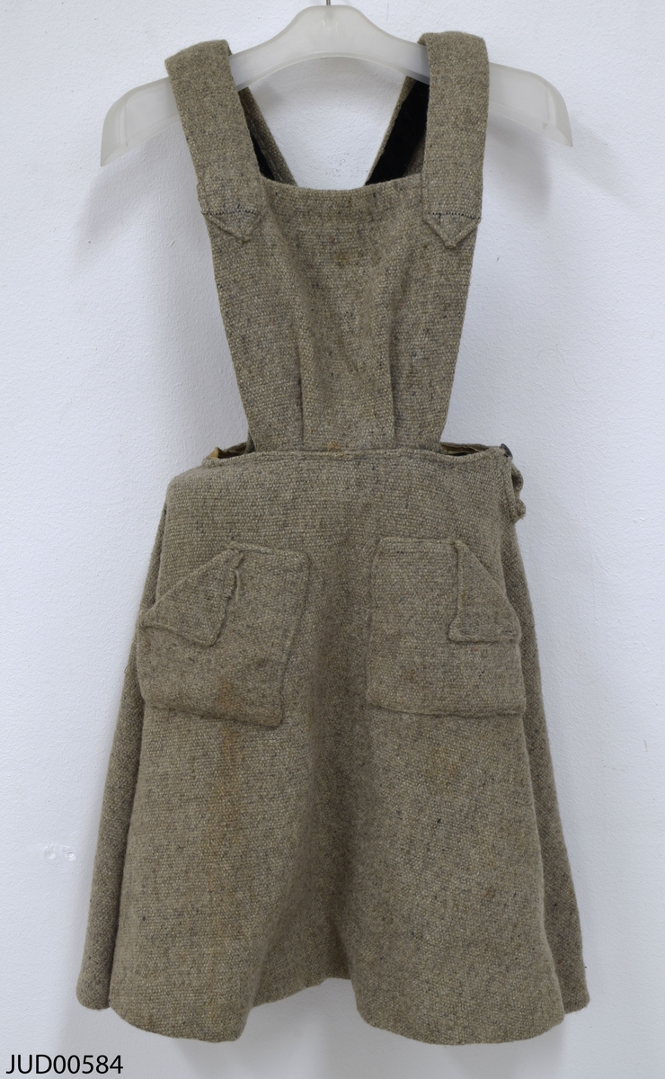 Grå klänning tillverkad av ett grovt tyg, troligtvis ull. Två fickor framtill och hängslen med knäppning i ryggen med knapp. Troligtvis sydd av en filt från koncentrationslägret Bergen-Belsen.