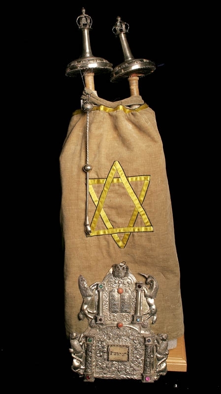 Torahsköld tillverkad av silver, med tillhörande pergamentbit med hebreisk text. Dekorerad med färgade stenar, och pelare flankerade med enhörningar och lejon. I mitten stentavlor med hebreisk text i.