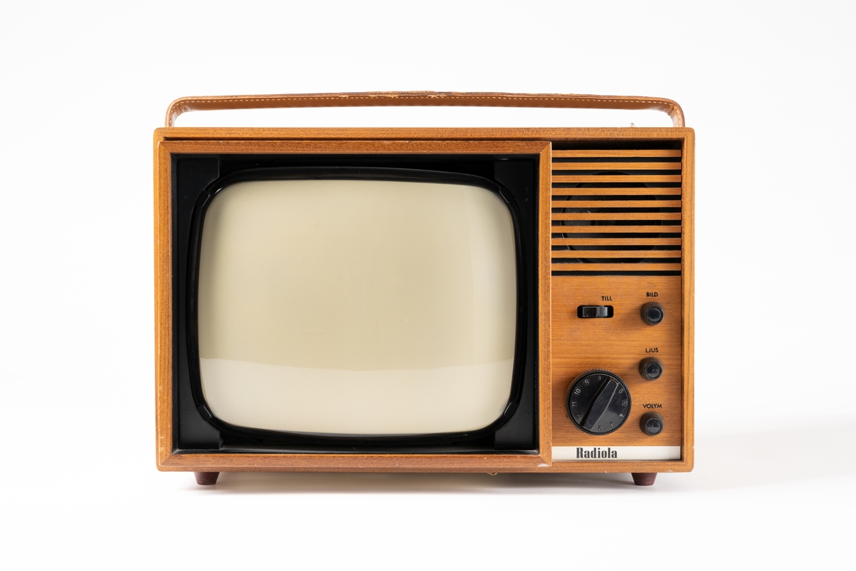 TV-apparat, Radiola Minett Typ 8410, av teak bärbar med handtag av läder. Upptill finns en uppdragbar antenn, baktill en vit sladd. På baksidan sitter en etikett med firmanamnet: "Karlsons RADIO TV FOTO I HUSKVARNA AB TEL. 30167-33910".