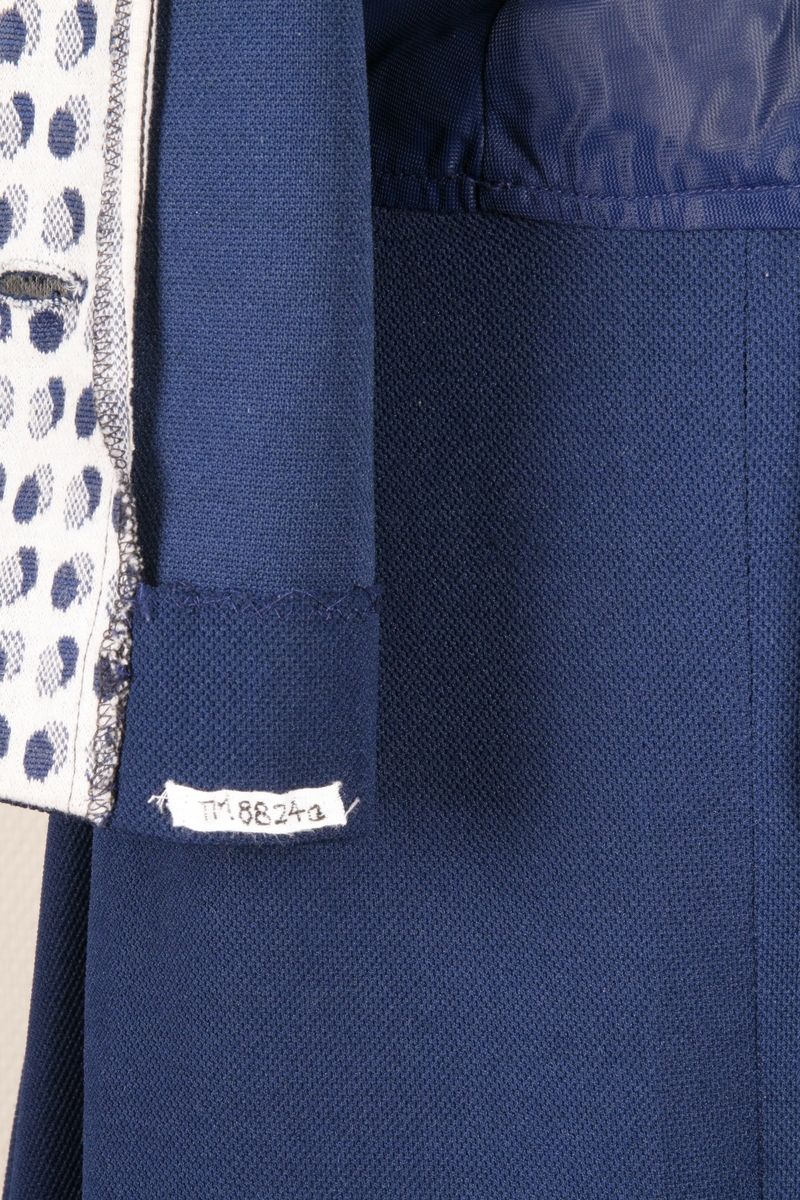 Mørk blå drakt med mønstret krage og ermkanter, jakke og skjørt. Fire knapper foran på jakken.