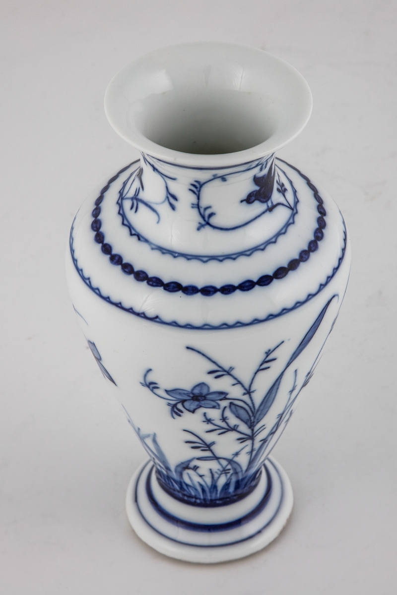 Vase i glasert porselen, dekorert med blomsterdekor i koboltblå underglasurmaling. Balusterformet koprus med utoverbrettet munningsrand, som hviler på en hevet fot.