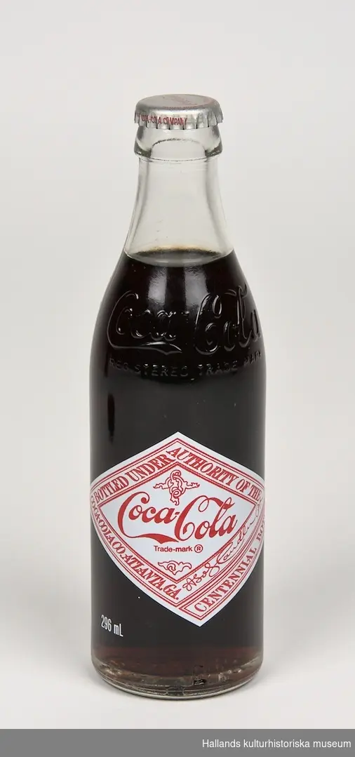 Läskedrycksflaska: Coca-Cola. Flaskan är tillverkad av klart glas och har två färgtryckta etiketter med text, varav en etikett har ett porträtt av John Pemberton - Coca-Colas grundare. I glaset finns två graveringar i form av Coca-Cola logotyper samt graverad text: "REGISTERED TRADE MARK". 296ml. Oöppnad.
