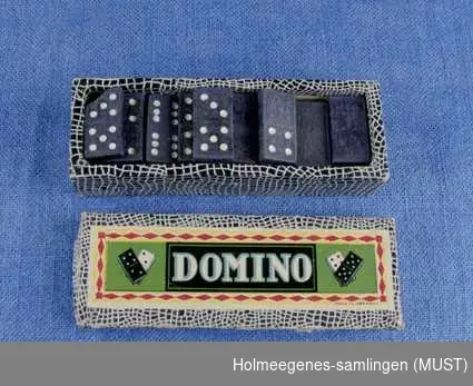 Esken er trukket med papir i svart og hvitt slangeskinnmønster. På lokket er et bilde av dominobrikker i hvitt og svart, med ordet DOMINO i midten.