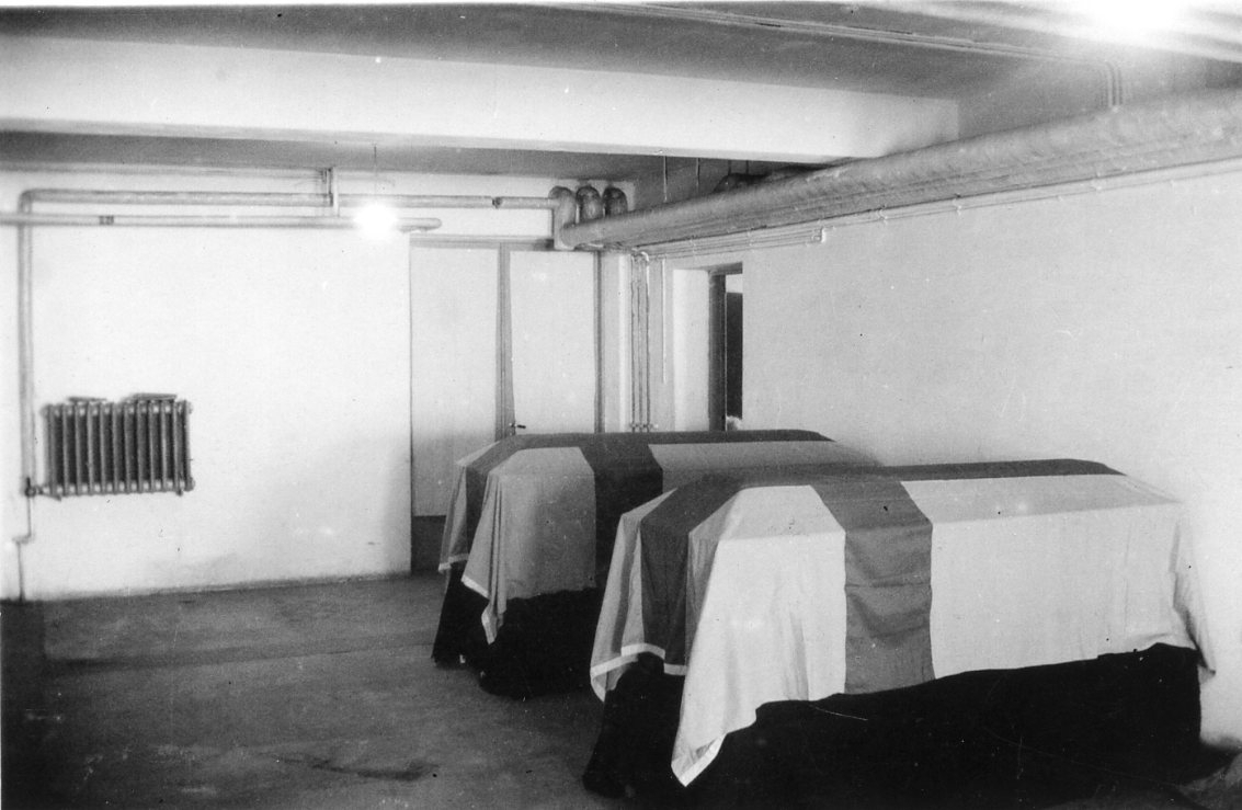 Två kistor draperade med svenska flaggan står i ett rum med ljusa väggar. Vattenburet element på väggen.