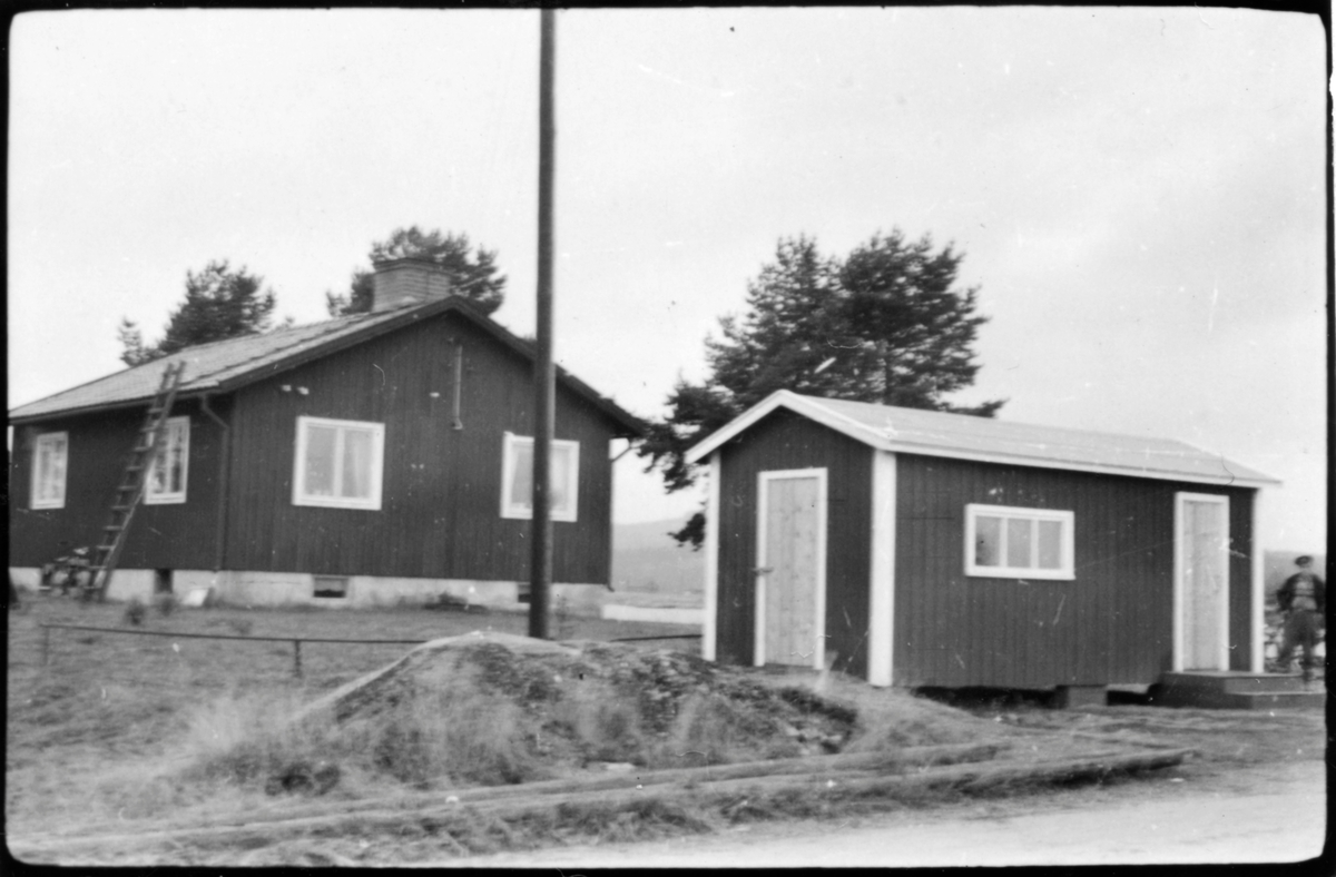 Vägstation AC 16, Åsele, färjvaktarbostad Gavsele. Bostadshus för färjvaktaren. I förgrunden mindre byggnad, eventuellt väntkur för resenärer. Mansperson till höger, troligen färjvaktaren. I bakgrunden färjleden.