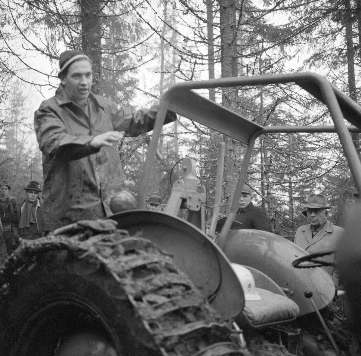 Fra en en driftsteknisk orientert skogdag i Strandbygda i Elverum (Hedmark) høsten 1963. Fotografiet viser en av instruktørene, en mann med regnjakke og topplue, som sto på trepunktkoplinga, eller kanskje på en lastebanke, bak førerplassen og gestikulerte mens han snakket til ei gruppe alvorlige, lyttende menn. Traktoren var utstyrt med halvbelter, som skulle øke bæreevnen, og den hadde veltebøyle. Hovedpersonen på dette bildet likner på Carl Fredrik Lindeman, som kom til Glommen skogeierforenings avdeling i Elverum som driftsteknisk konsulent i 1961. Ti år seinere fikk han tittelen skogbrukssjef.  Mannen vi ser deler av ansiktet til bak verltebøyla på traktoren er skogeier Rolf Sætersmoen fra Elverum.