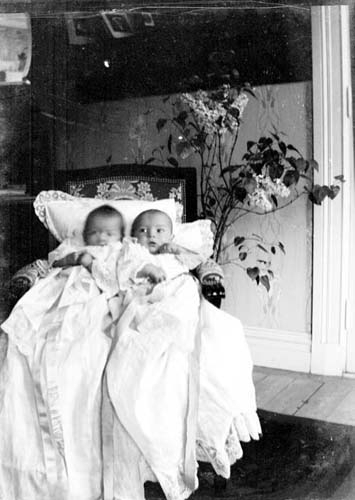 Två små barn halvsitter i en klädd stol. De är klädda i vad som ser ut som dopkläder.