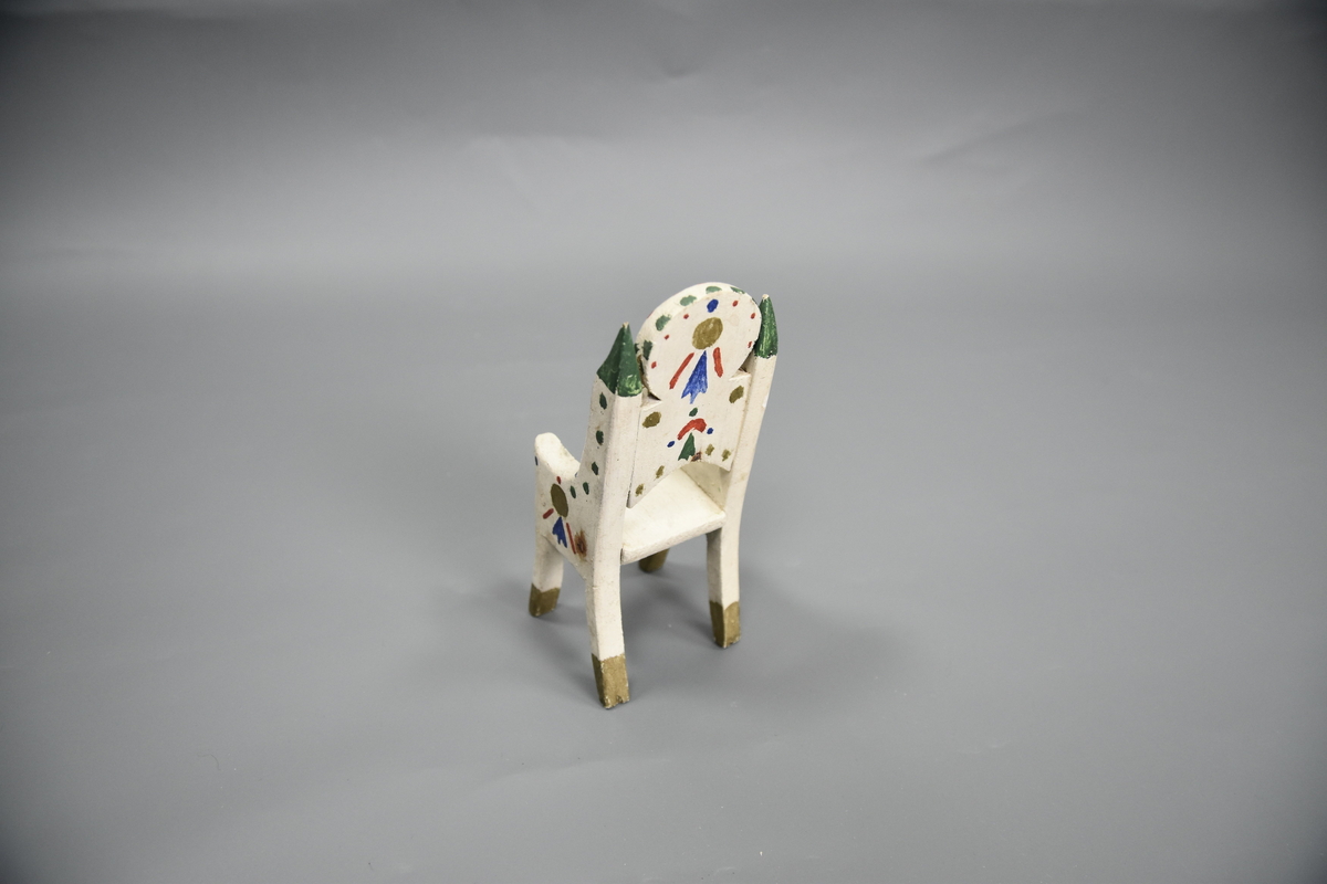 Stol til dokkemøblement i fem deler. Denne stolen er litt mindre enn de tre andre. Møblene er hvitmalte med dekor i ulike farger. 
Møblementet er fra givers tidlige barndom, muligens 1928-1930. Møblene var til hennes første dokkestue, bygget av far av en appelsinkasse. 