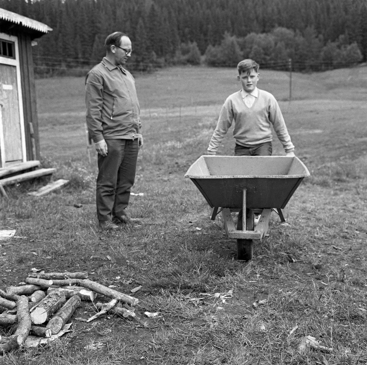 Gutt med trillebår, fotografert under en 4H-leir i Søre Osen i Trysil i Hedmark i 1962. Da dette bildet ble tatt sto jenta med bøyde knær, enten for å løfte eller sette fra seg den tunge båra. En voksen mann sto og betraktet henne, antakelig for å bedømme arbeidet. Bak ham ser vi en utedo.