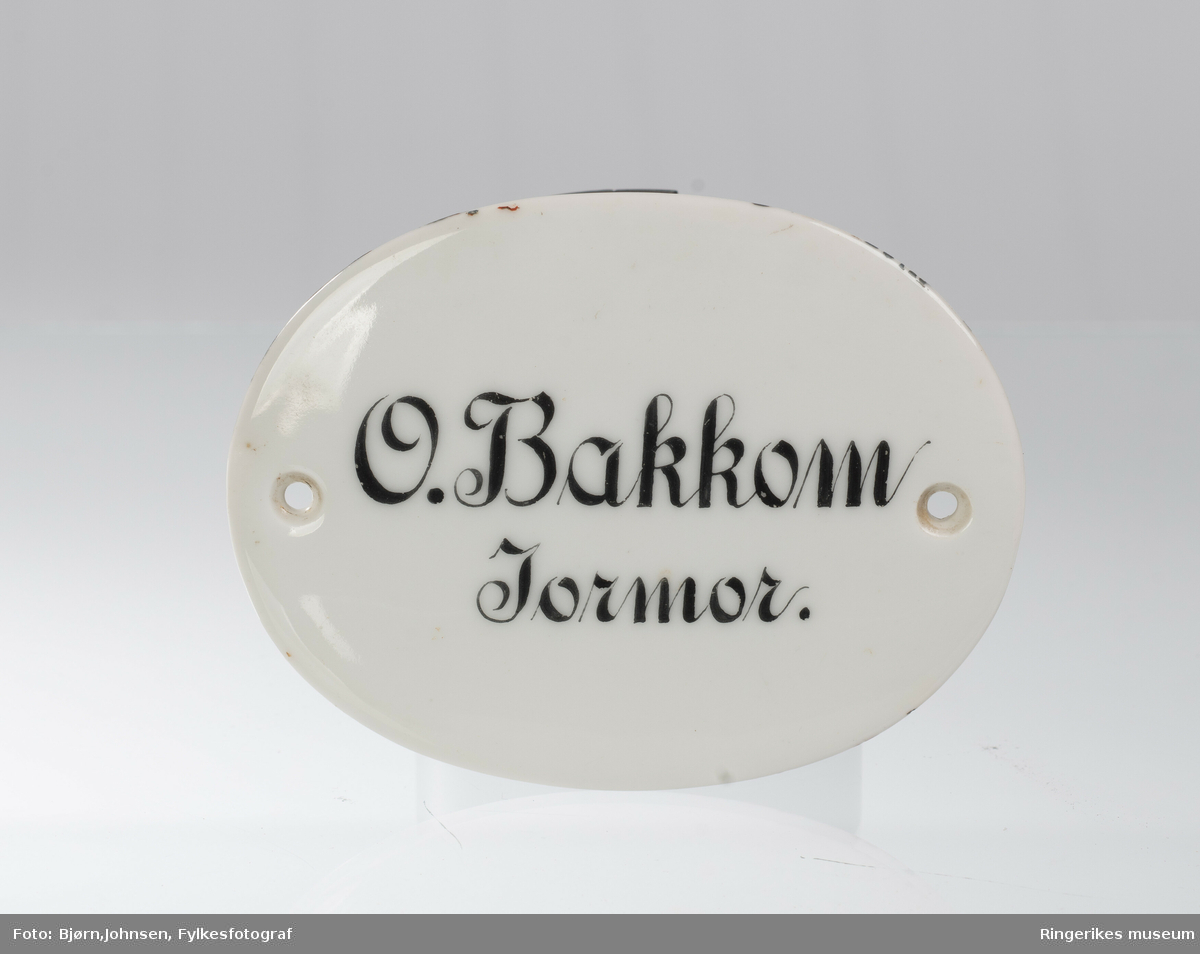 Oval hvitglassert navneskilt i keramikk. Svart skrift: O. Bakkom. Jormor. Hull til feste i begge sider.