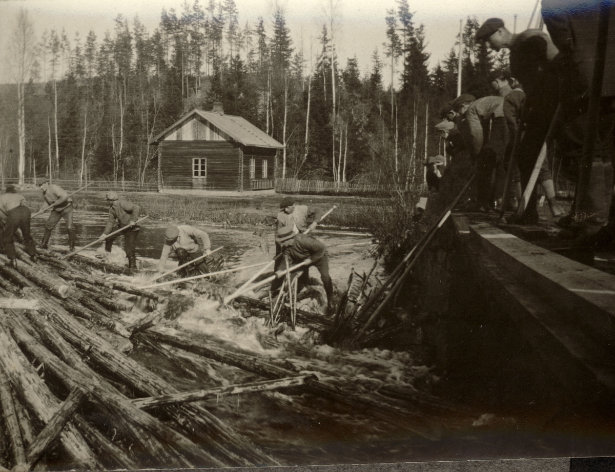 En gjeng tømmerfløtere arbeider med fløting på og ved Knappen bru over Juråa i Nord-Odal. Et tømmerhus står på den andre siden av elva. Fotografert 1921.