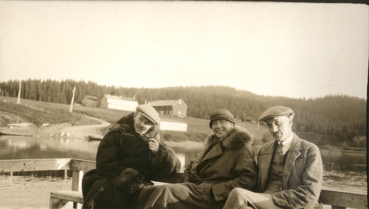 Harald Platou, Mia og Oscar Collett sitter på en ferge over Namsen i Trøndelag. I bakgrunnen et stort gårdstun. Fotografert september 1923 i forbindelse med elgjakten.