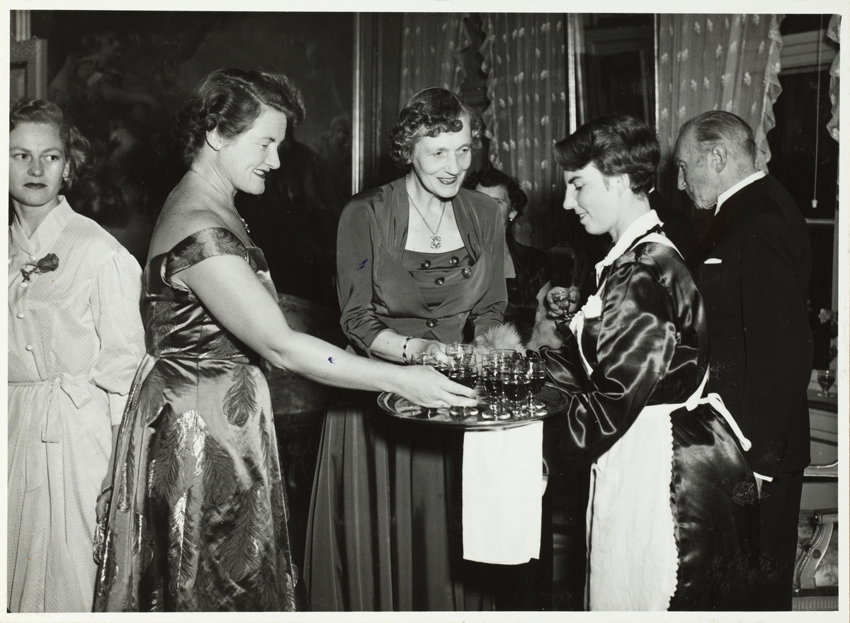 To kvinner i selskapskjoler får servert drinks av kvinnelig servitør. En av kvinnene er Ingeborg Poulsson, gift med arkitekt Esben Poulsson. Anledningen er fest på Bogstad gård for Arkitektforeningen. Fotografert i september 1953.