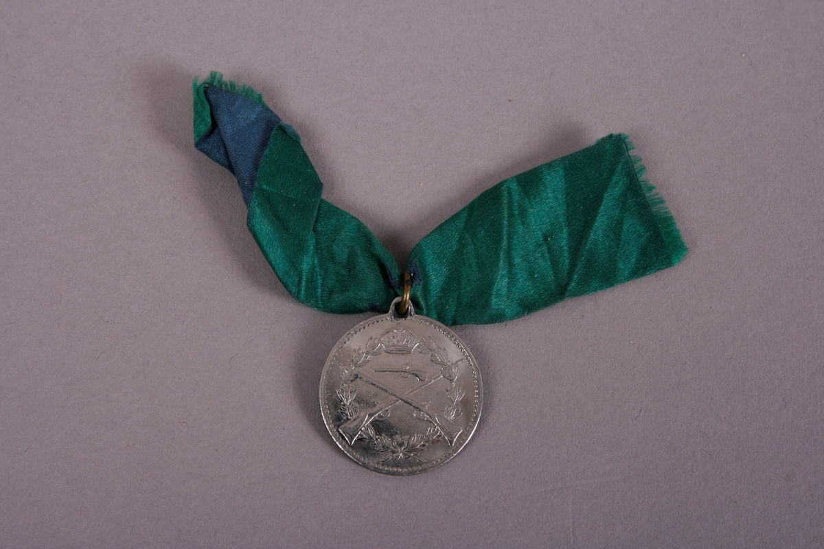 Medalje med grønt bånd, festet i ring på toppen.