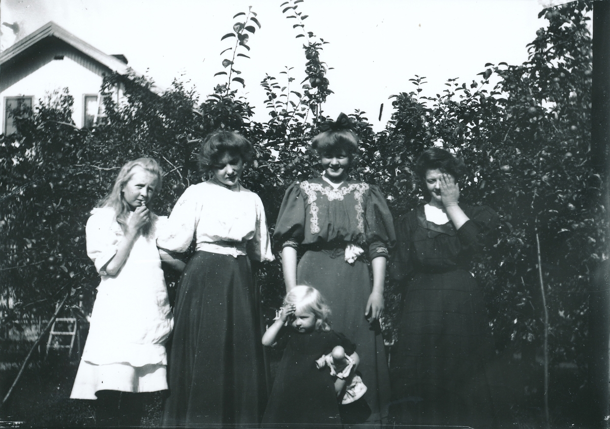Gruppebildene av kvinnene i Rydgrenfamilien i hagen utenfor bestyrervillaen på Kistefos. Kvinnene har oppsatt hår og lange kjoler med vide ermer til albuene. Den minste jenta holder en dukke. Arbeiderboligen, Boligen, i bakgrunnen.