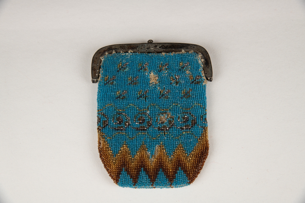 Liten pung, med avrundede nedre hjørner, med  små perler i egyptisk blått og gyllent, og mørkebrunt, siksakbord nederst. På spennen   inngravert  :   E J C L den 26 Februar 1841