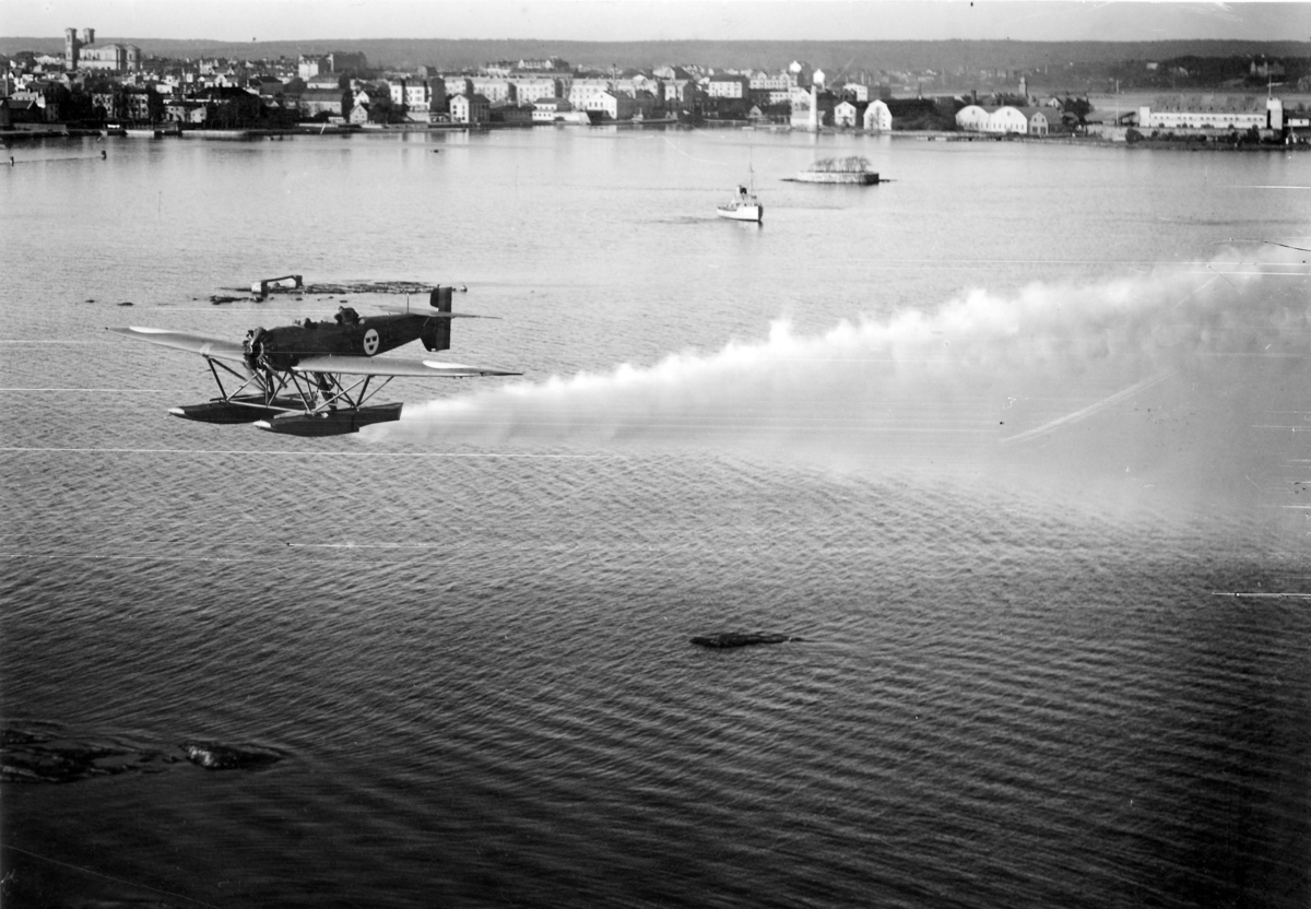 Dimbildning med flygplan S 5 Heinkel He 5 Hansa, nummer 2452 över vatten vid Karlskrona. Stadsbild i bakgrunden. 1930-tal.