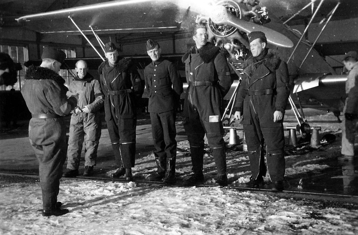 Ordergivning i hangarport framför flygplan S 5 Heinkel He 5 Hansa. En militär i flygoverall föredrar inför flygning framför fem andra militärflygare i en hangarport. Vintertid, 1930-tal.