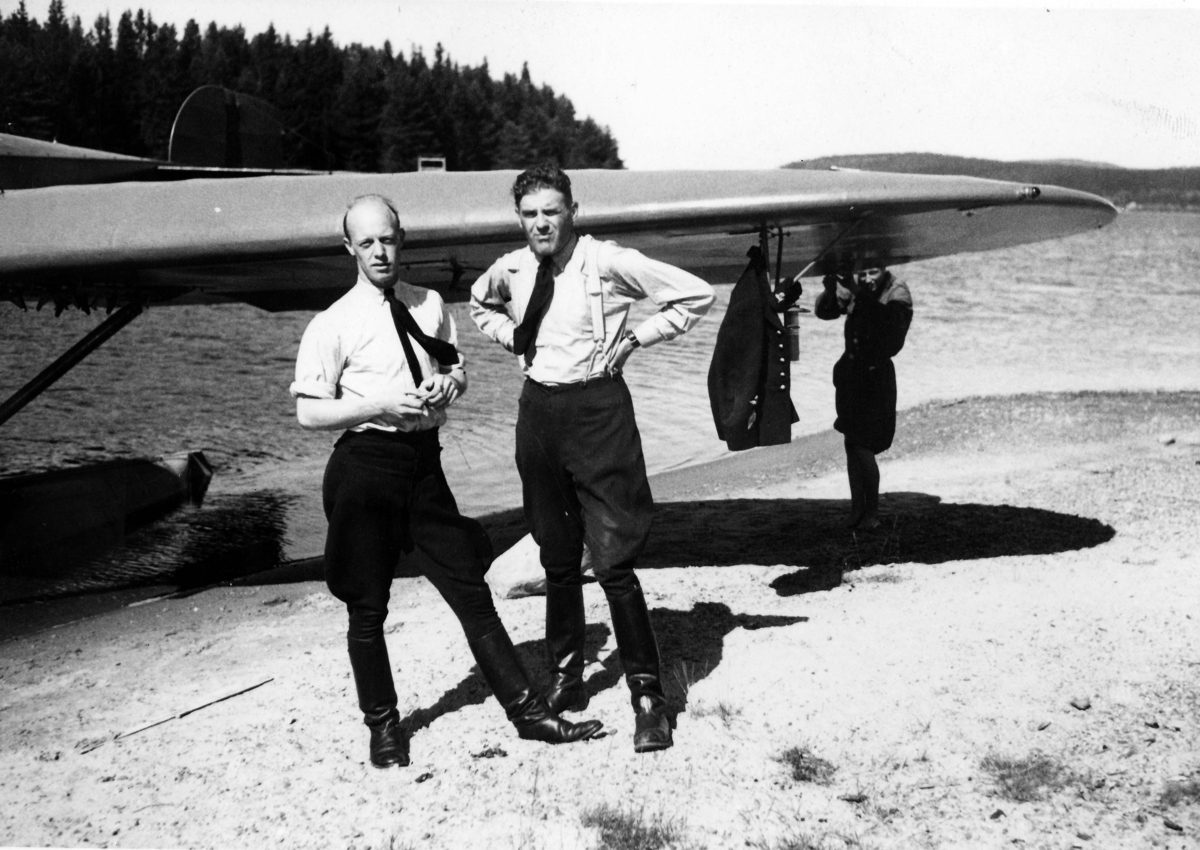 Två män står framför flygplan S 5, Heinkel He 5 Hansa på en strand. Mannen till vänster är militäre flygaren Verner Tornberg. I bakgrunden arbetar en mekaniker med flygplanets vinge. 1930-tal.