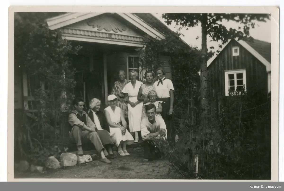 Svart vitt fotografi, grupporträtt. Kvinnor och män sitter respektive står framför ett hus.