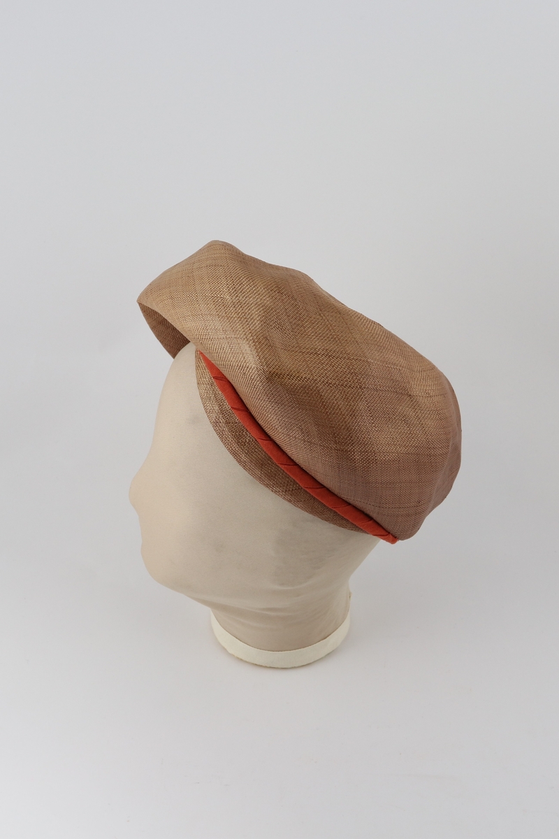 Brun hatt med brettet brem forann og påsydd remse med tvinnet tekstil ved bretten.