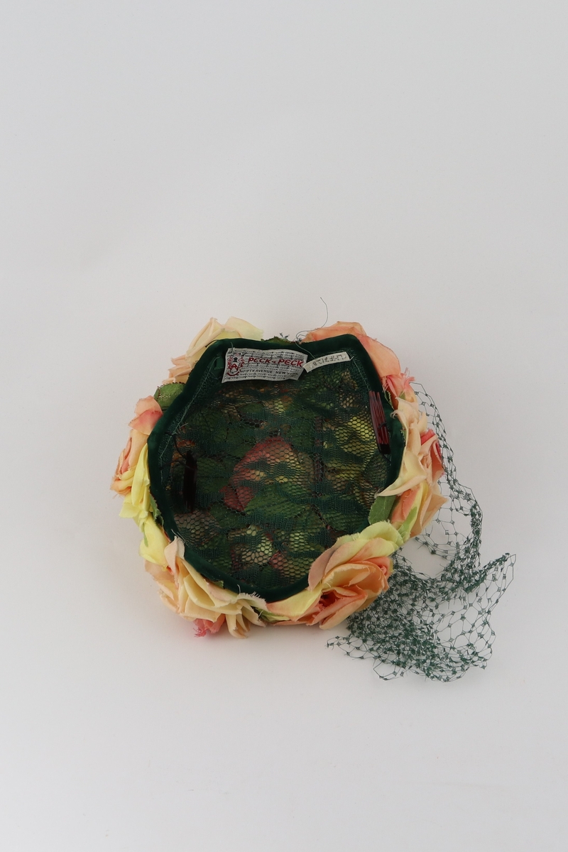 Hatt av stiv grønn netting dekket av gule og rosa roser av tekstil. Netting festet med tråd i et punt på bakre del av toppen, sprer seg herfra til å dekke den ene siden og et nytt fest på fronten. En spenne på hver side for å feste hatten til hodet.