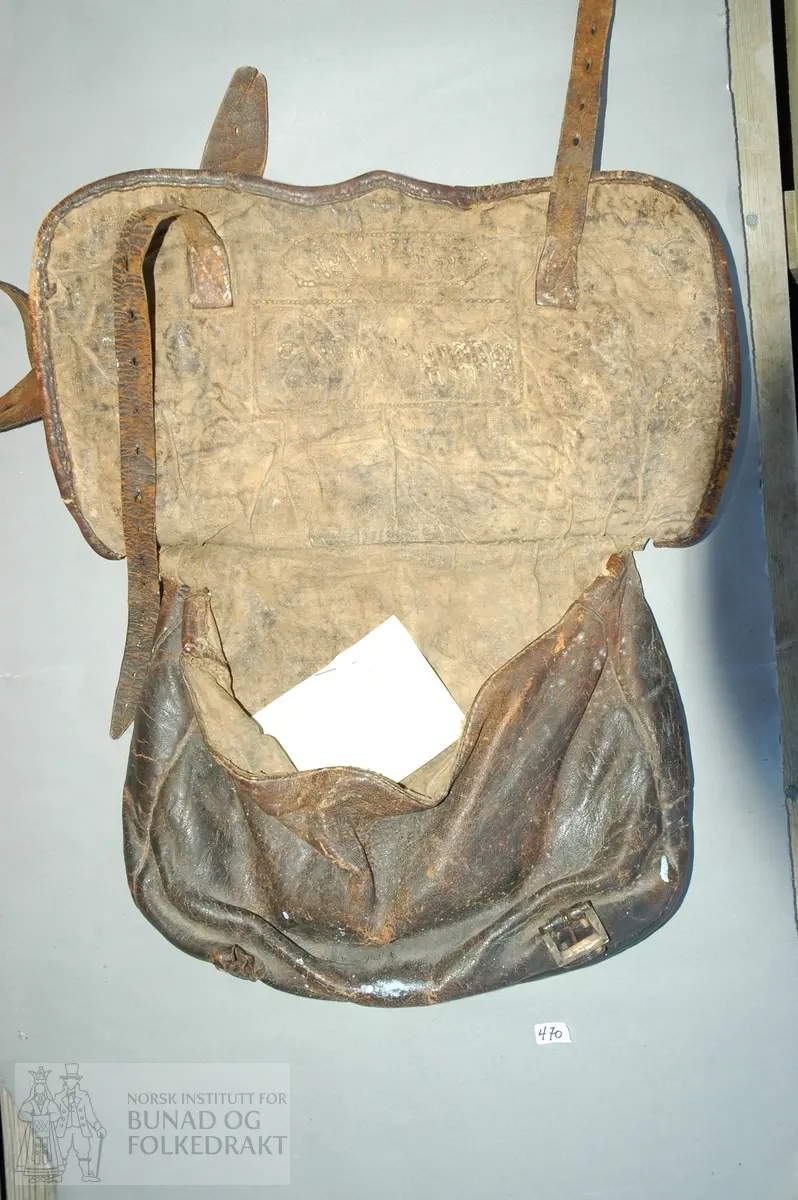 Taske med lokk av mørk brunt skinn. Lokket snøres med to reimer. Skulderreim. Påsydd på lokket: Ingulf Mitdahl 15/4 . 80