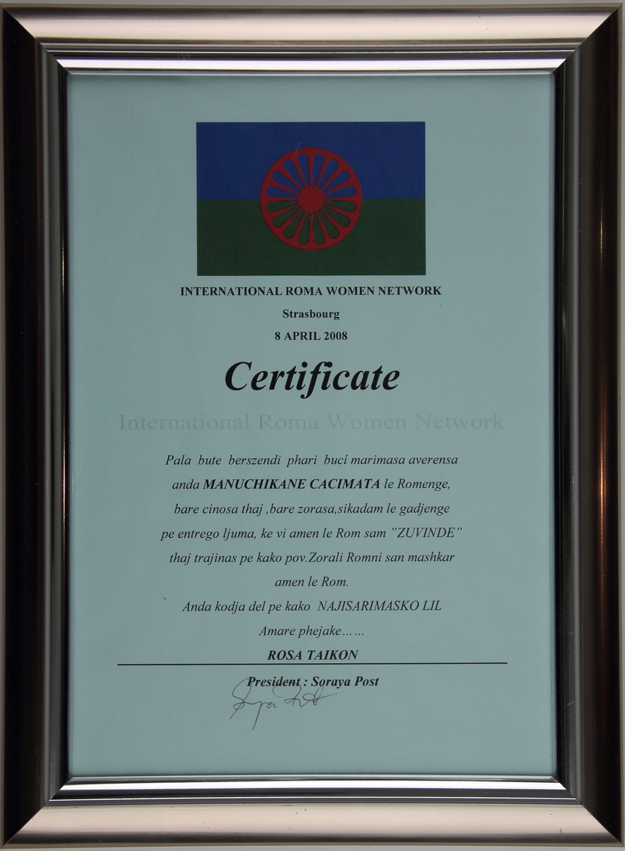 Ett certifikat från International Roma Women Network till Rosa Taikon. Certifikatet är inramat i en metallisk ram och överst finns den romska flaggan.