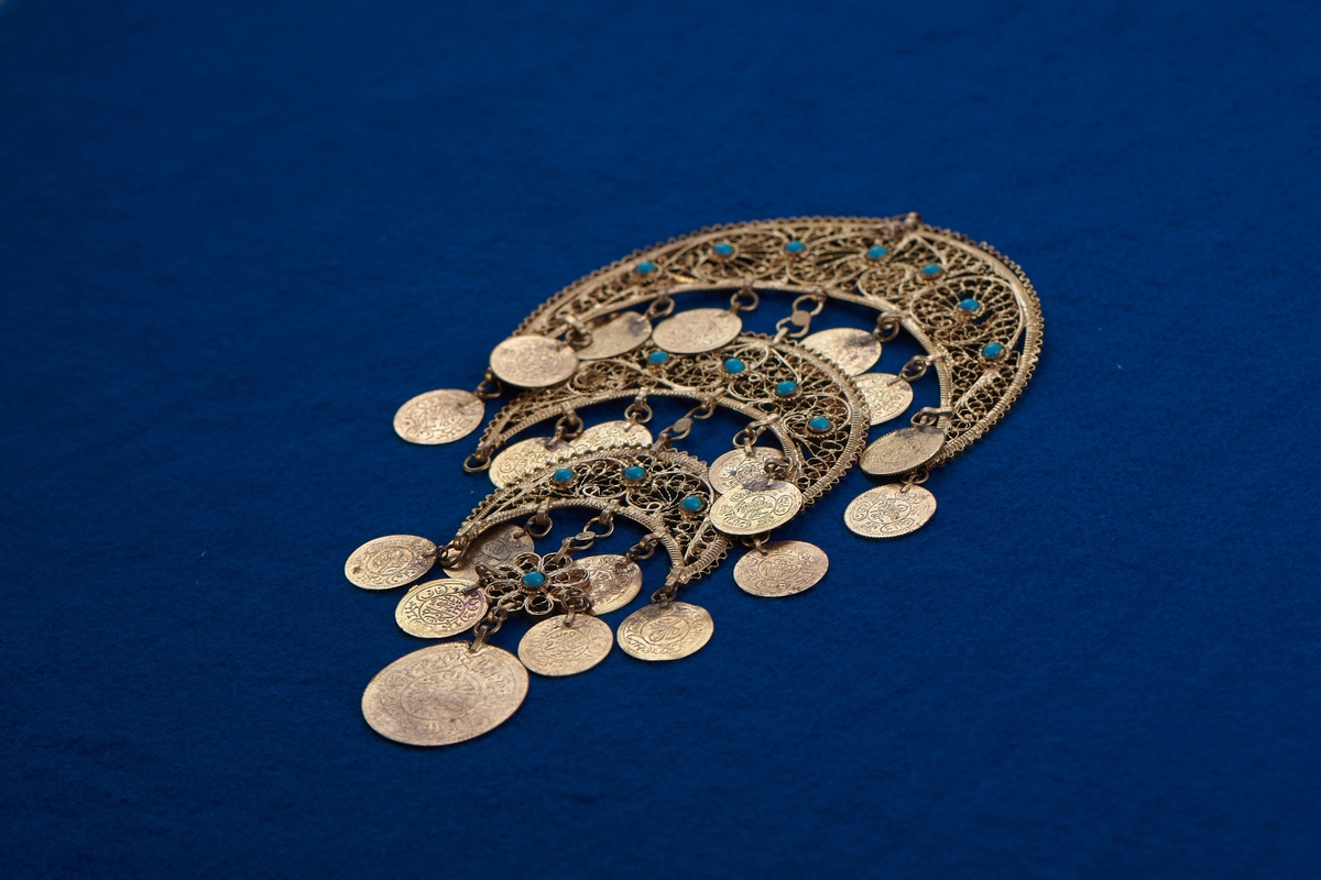 Del av halssmycke, bestående av metallbitar i form av tre halvmånar. Smycket är skapat i gul metall med infattade turkosfärgade stenar. Från varje halvmåne hänger cirklar i bindöglor. Smycket är troligen inköpt av Rosa Taikon vid en resa utomlands, och hängde senare på anslagstavlan i hennes ateljé.