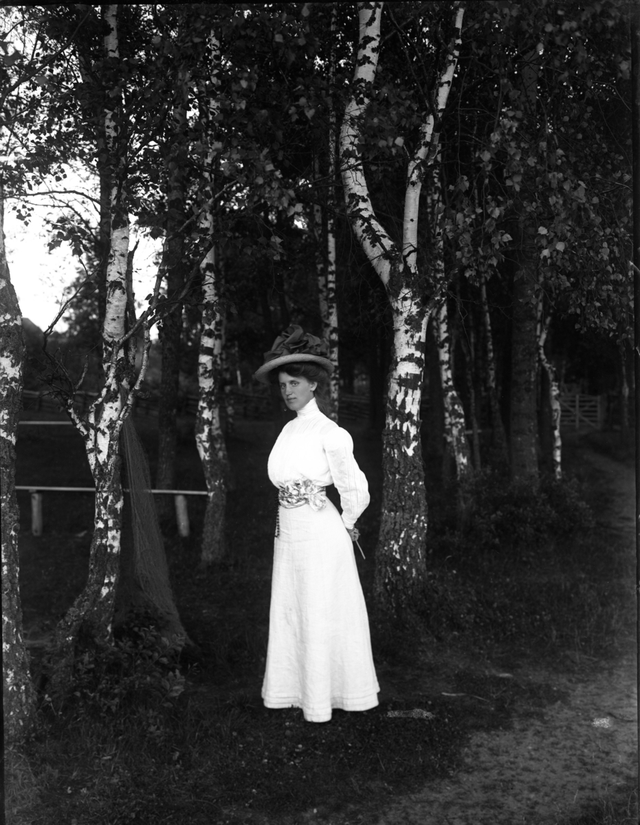 En ung kvinna i ljust och stor hatt står vid en björkdunge intill en stig. Det är modisten Edit Eriksson från Jönköping.
