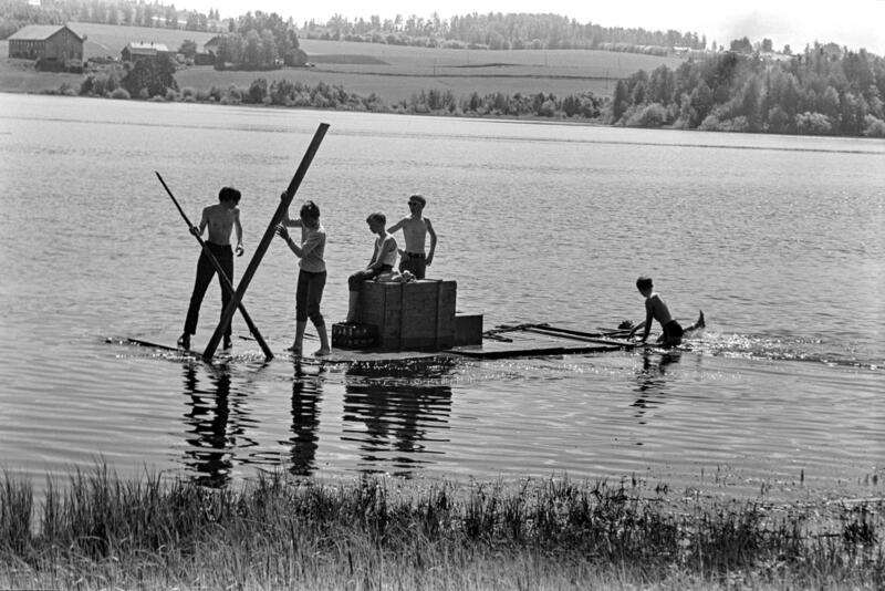 Svart-hvitt bilde av fem ungdommer i shorts som seiler på en hjemmelaget flåte.