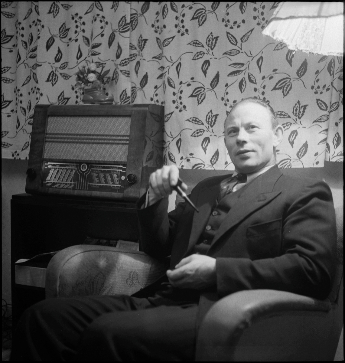 En mann sitter i en godstol i ei stue og røyker pipe. Det er en stålampe bak han og en radio på siden.