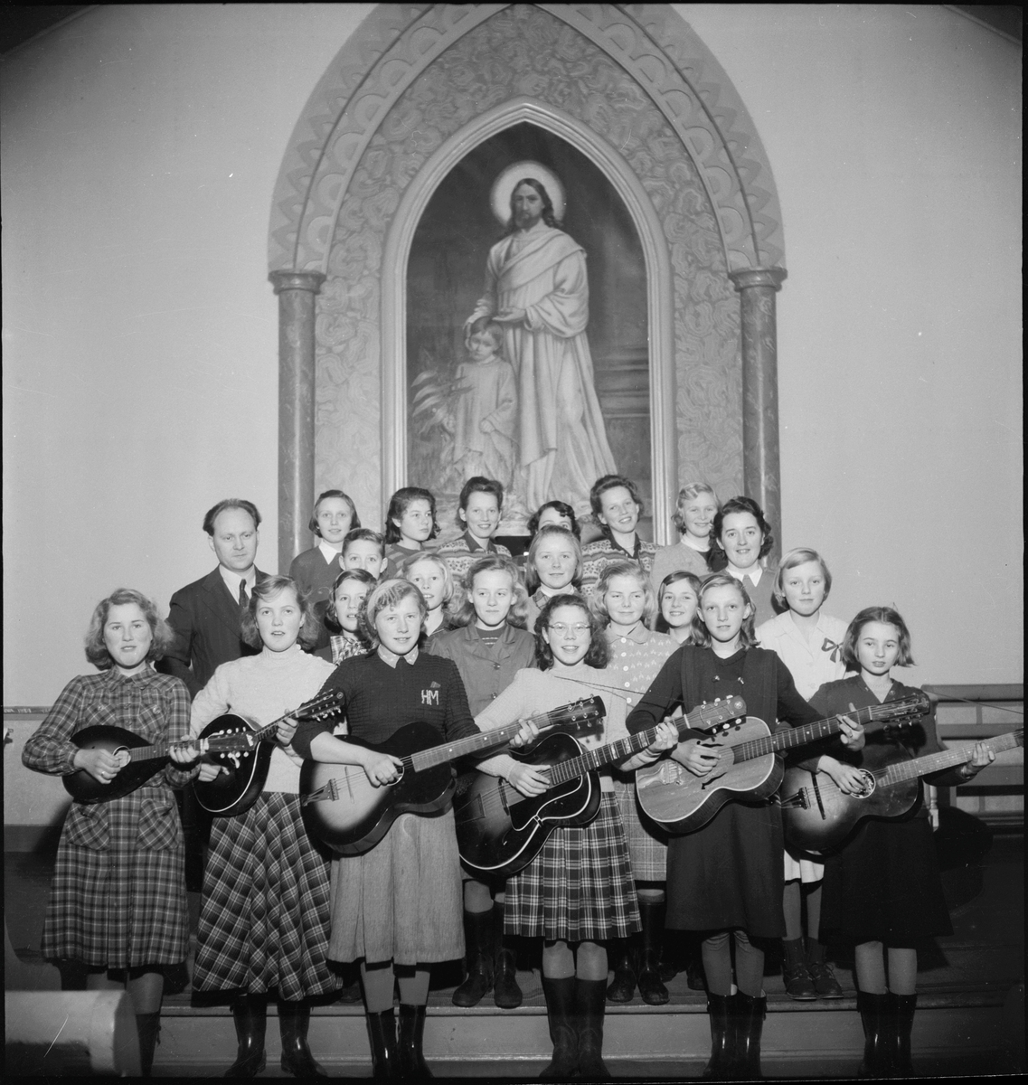 En  jentegruppe foran altertavlen i Metodistkirken. Flere av jentene har gitar eller ukulele.