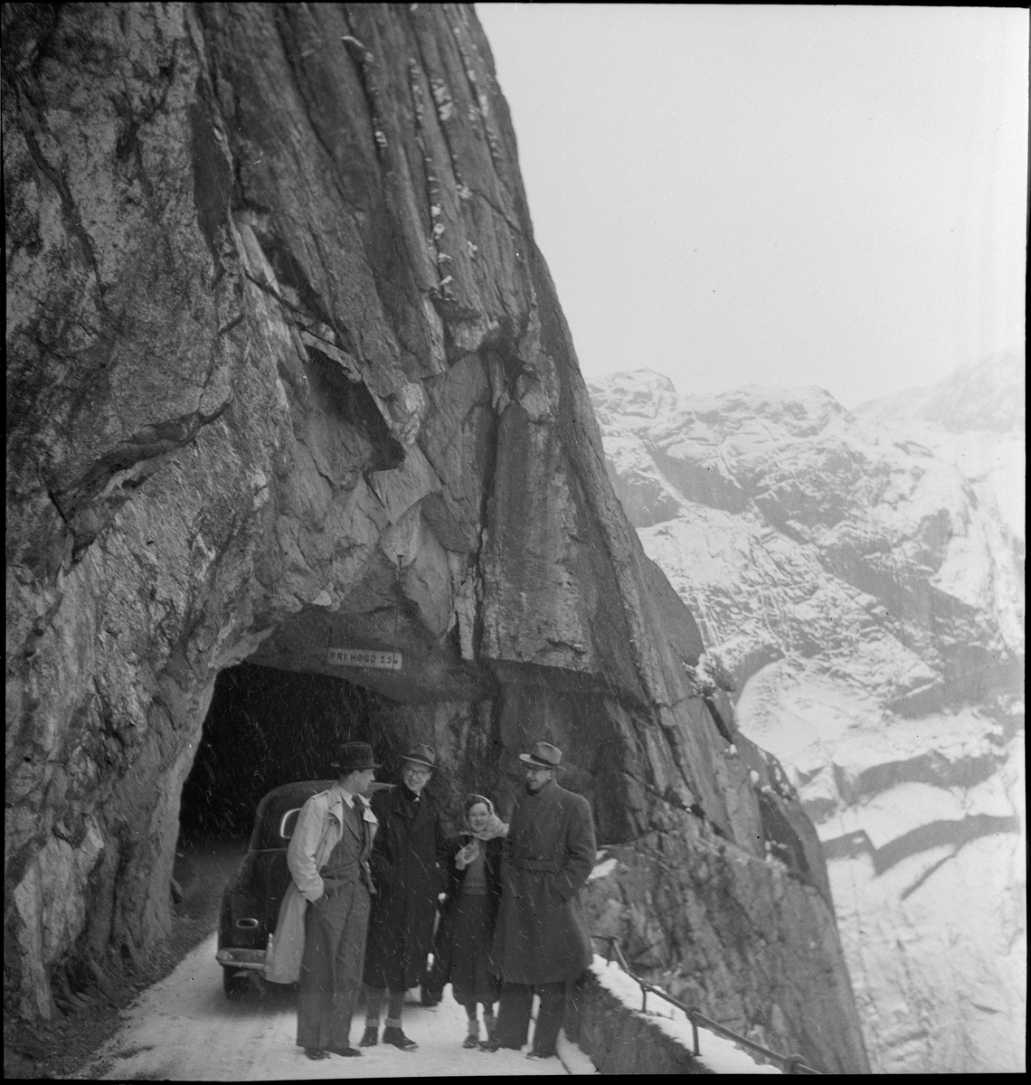 Tre menn og en kvinne på biltur en vinterdag. Det er bratte fjellsider med en tunell og en dal med en fabrikk i enden av et vann.