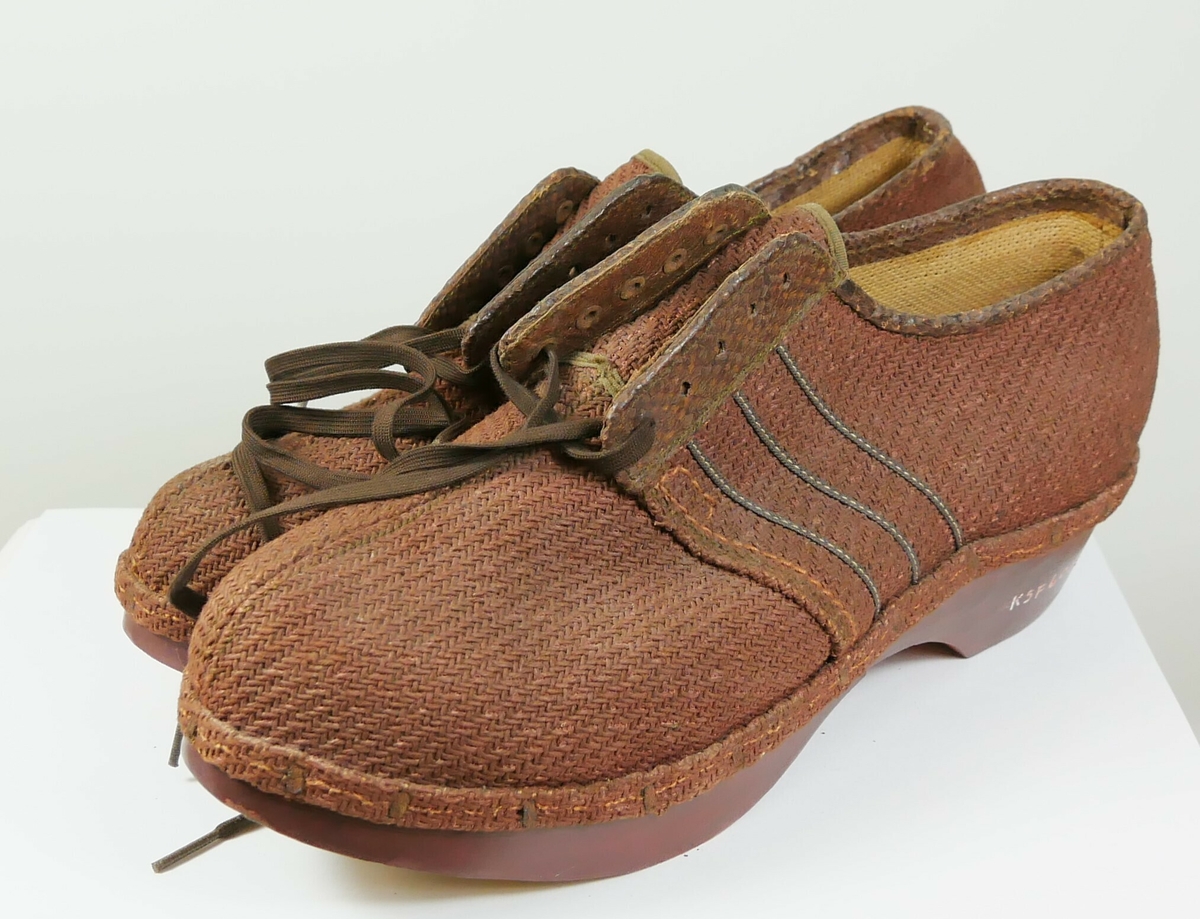 Røde sko av tekstil som er naglet til en en rødmalt såle av tre Under sålen er det ni gummiknotter. Det er brune lisser til knyting foran på skoen. 