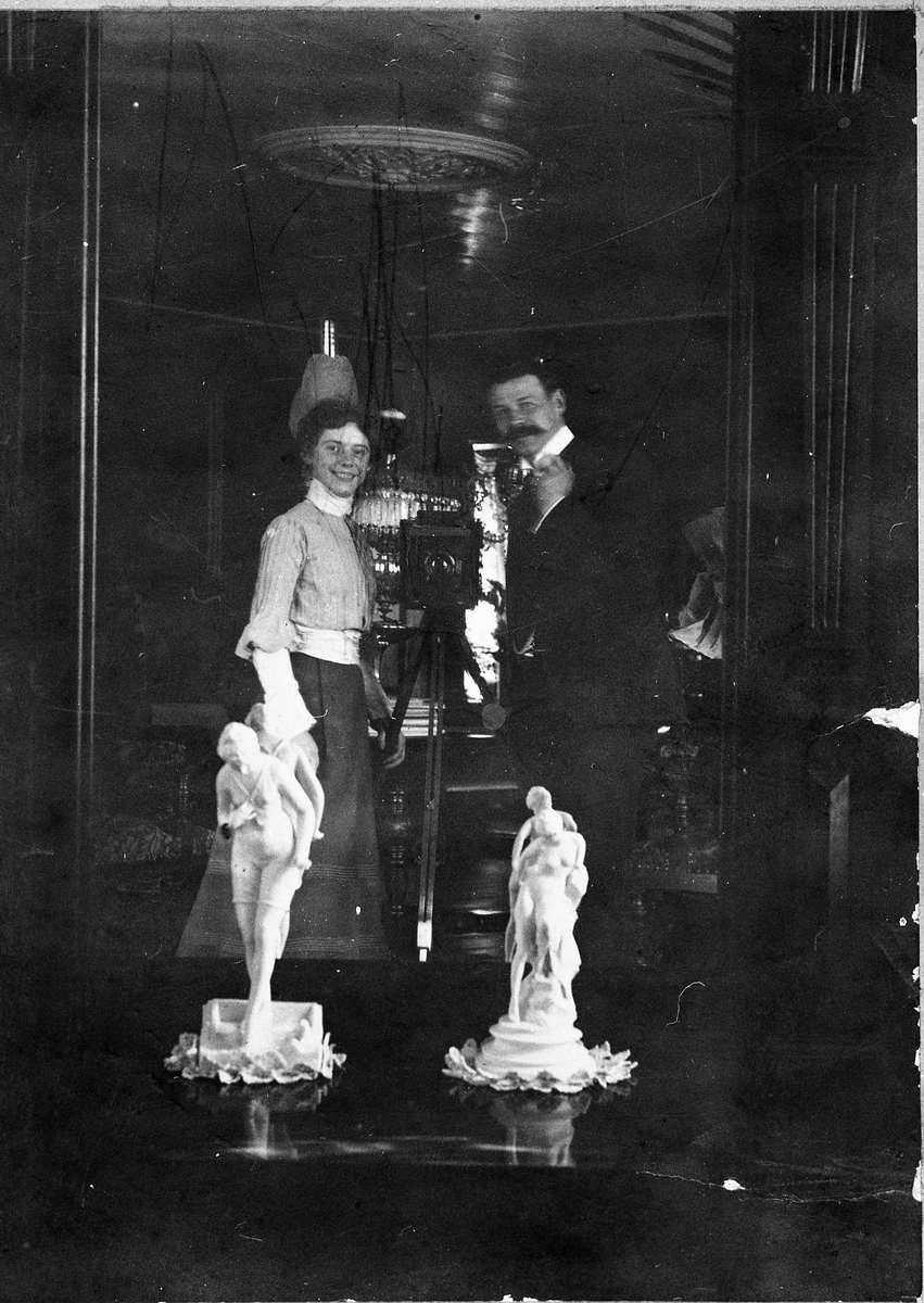 Bergljot Rogneby (g.Holm) og Gustav Mikkelsen avbildet foran det store speilet i salen på gården Rogneby, Østre Toten, 1905. Mellom de to står et fotoapparat. Mikkelsen har tatt bildet med selvutløser.