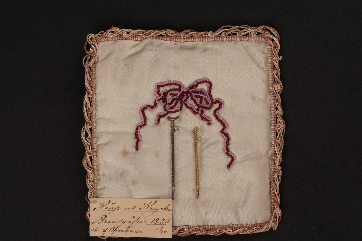 Käpp av guld och krycka av silver, uppfästa på vitt siden med pärlbroderier. 
Sidenet är i två lager med fyllning. Kanten är dekorerad med sidentråd i vitt och brunt. På framsidan ovanför kryckan och käppen finns en pärlbroderad rosett i två färger, vinrött och rosa.
På framsidan sitter en fastsydd etikett med följande text:
"Käpp och Krycka Brudgåfva 1820 sk. af Öfverstinnan Örn".