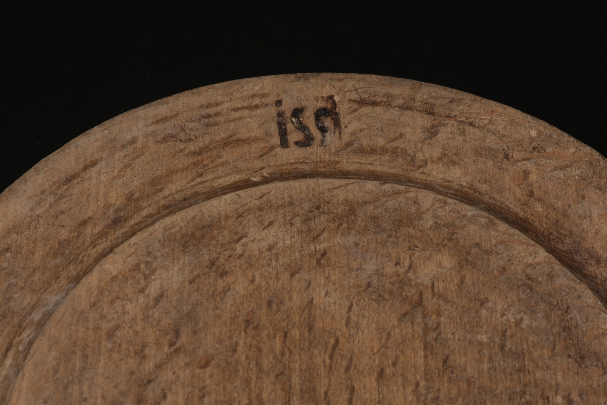 Rund, flat, svarvad trädisk med bräm.
På undersida finns en brännstämpel med initialerna ISS.