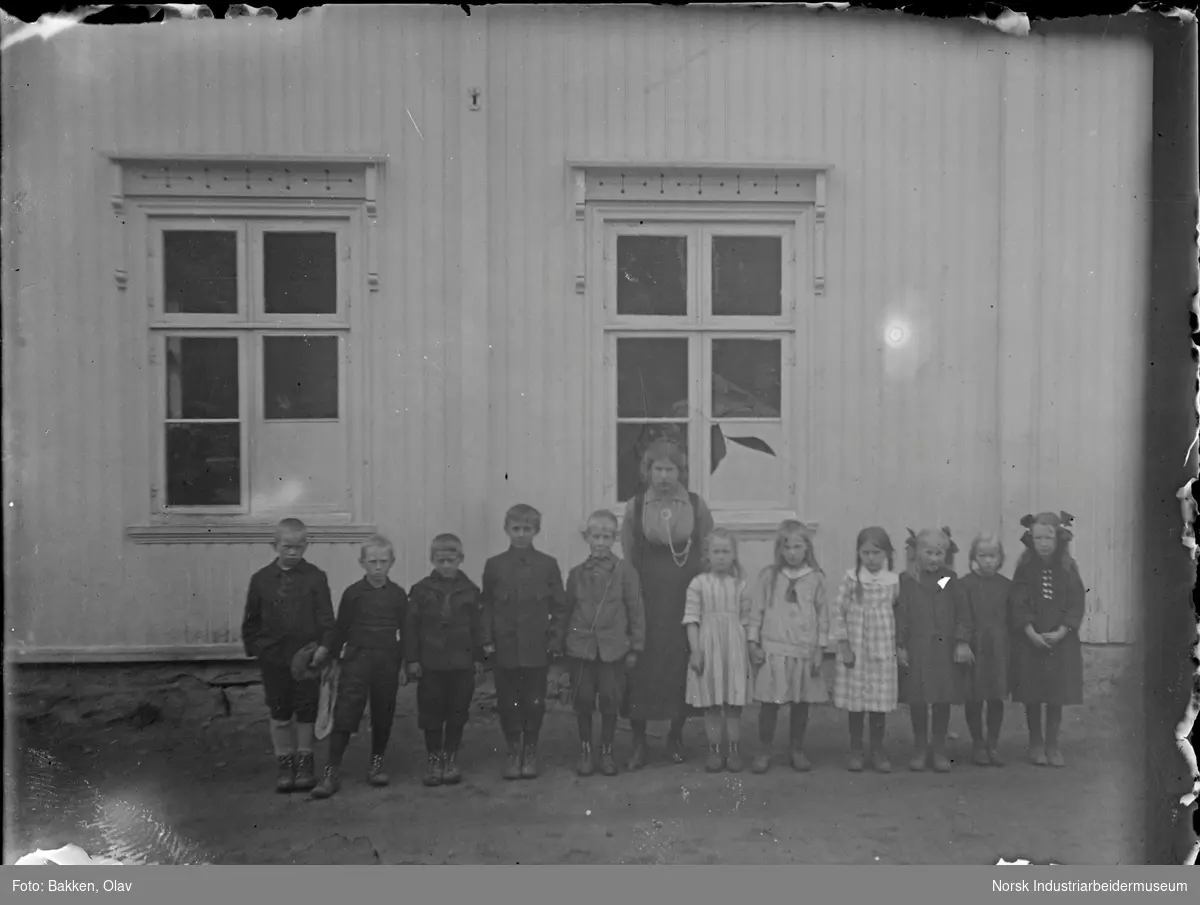 Skoleklasse på Klokkargarden skole. 11 elever og en lærer oppstilt foran skolebygning. Jentene står på lærerens venstre side, guttene på lærerens høyre side.