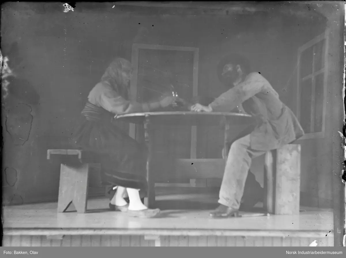 Amatørteater. To mennesker spiller skuespill sittende ved et bord på scene.