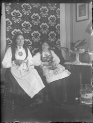 To jenter sittende i stue i bunad med håndarbeid i hendene. 