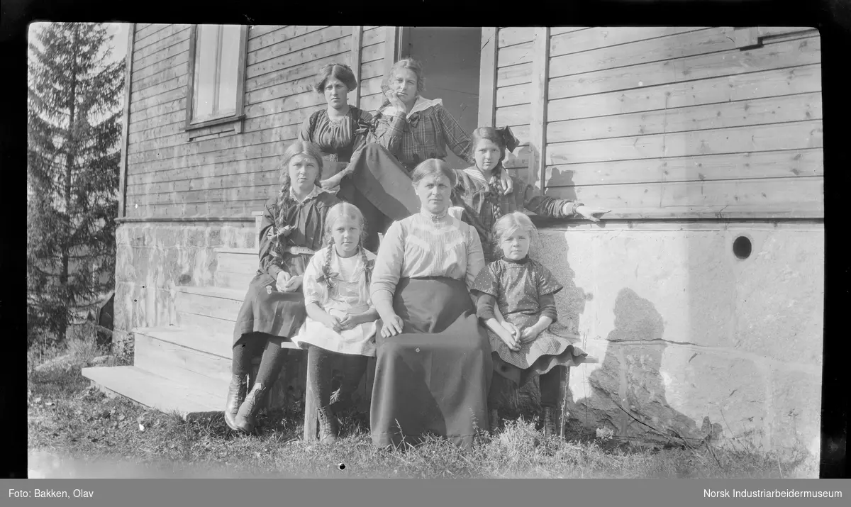 Kvinner og barn sittende på trapp foran bolighus. Jentene med fletter i håret og sløyfer.