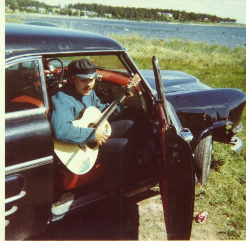 Ung mann sitter i døråpningen av bil og spiller gitar.