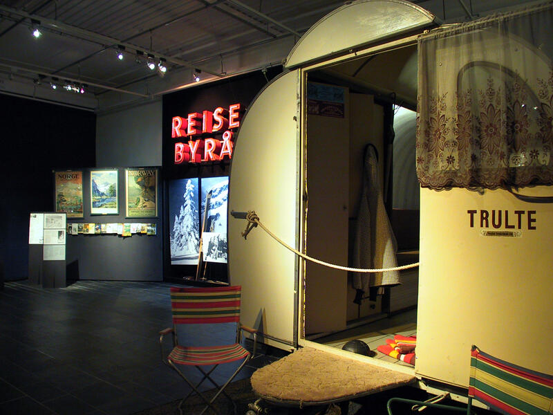 Oversiktsbilde fra utstillingen Hilsen fra Norge - Utstilling om turisme. Utsnitt av en campingvogn, vegg hvor det står Reisebyrå samt en vegg med noen bilder.