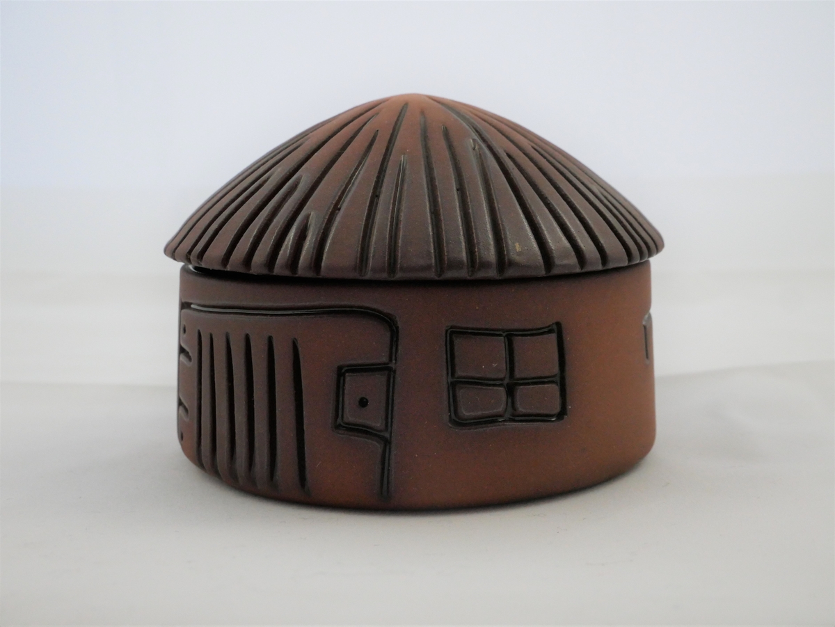 Lita krukke i keramikk, med lok. Rundt tverrsnitt. Korpus er sylinderforma, medan loket er forma som ei kjegle. Krukka førestiller eit hus, og har innskorne detaljar.