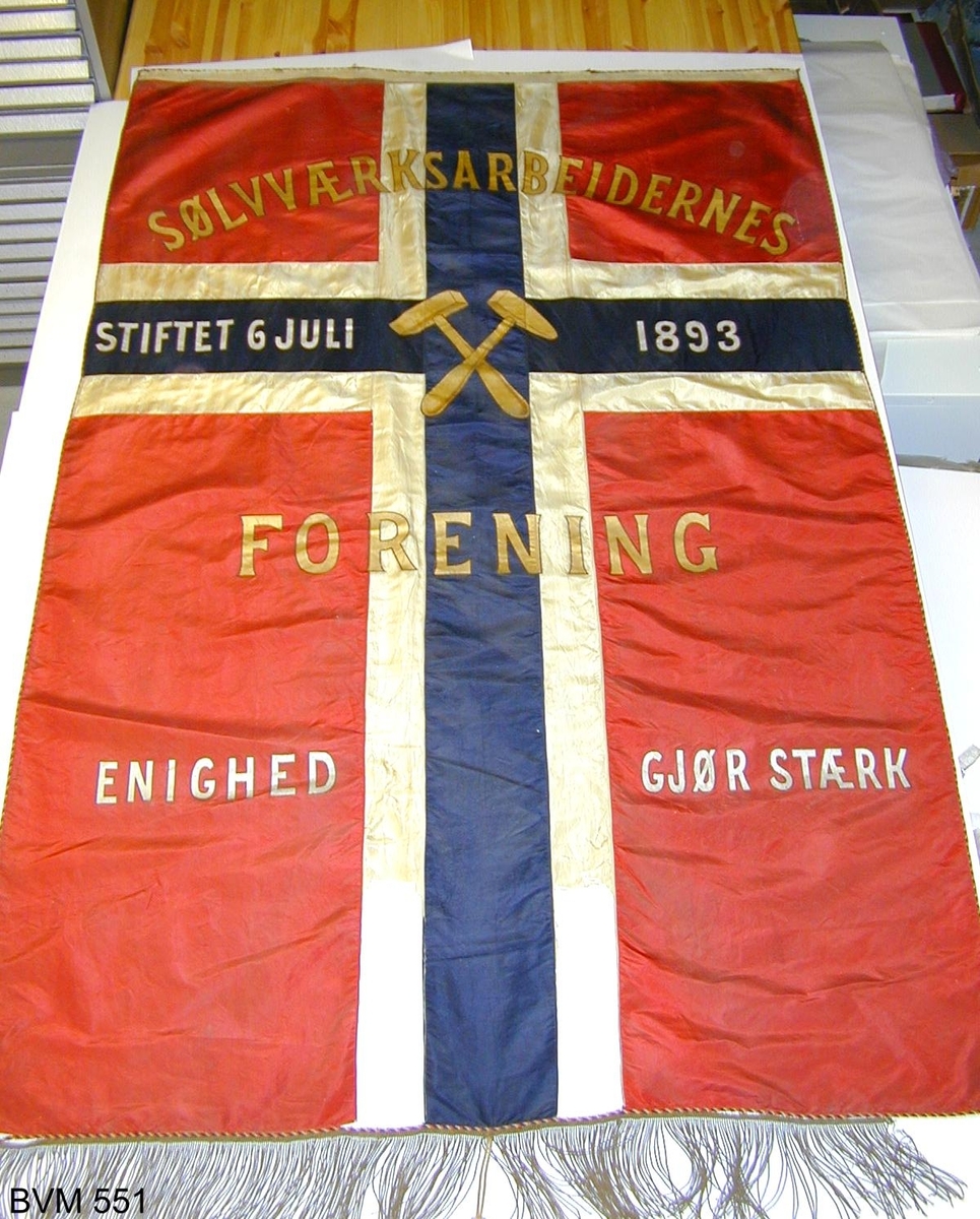 Sølvverksarbeidernes fagforeningsfane fra 1893. Fanen er et norsk flagg med tre "gulldusker" og frynser nederst. På fanen står det: "SØLVVÆRKSARBEIDERNES FORENING STIFTET 6 JULI 1893 ENIGHED GJØR STÆRK". Mellom dato og årstall står korslagt hammer og bergsjern. Fanen er generelt slitt og har to store hull i nedre del der det kvite i flagget er borte. I tillegg er frynsene løse, spesielt på høyre side.